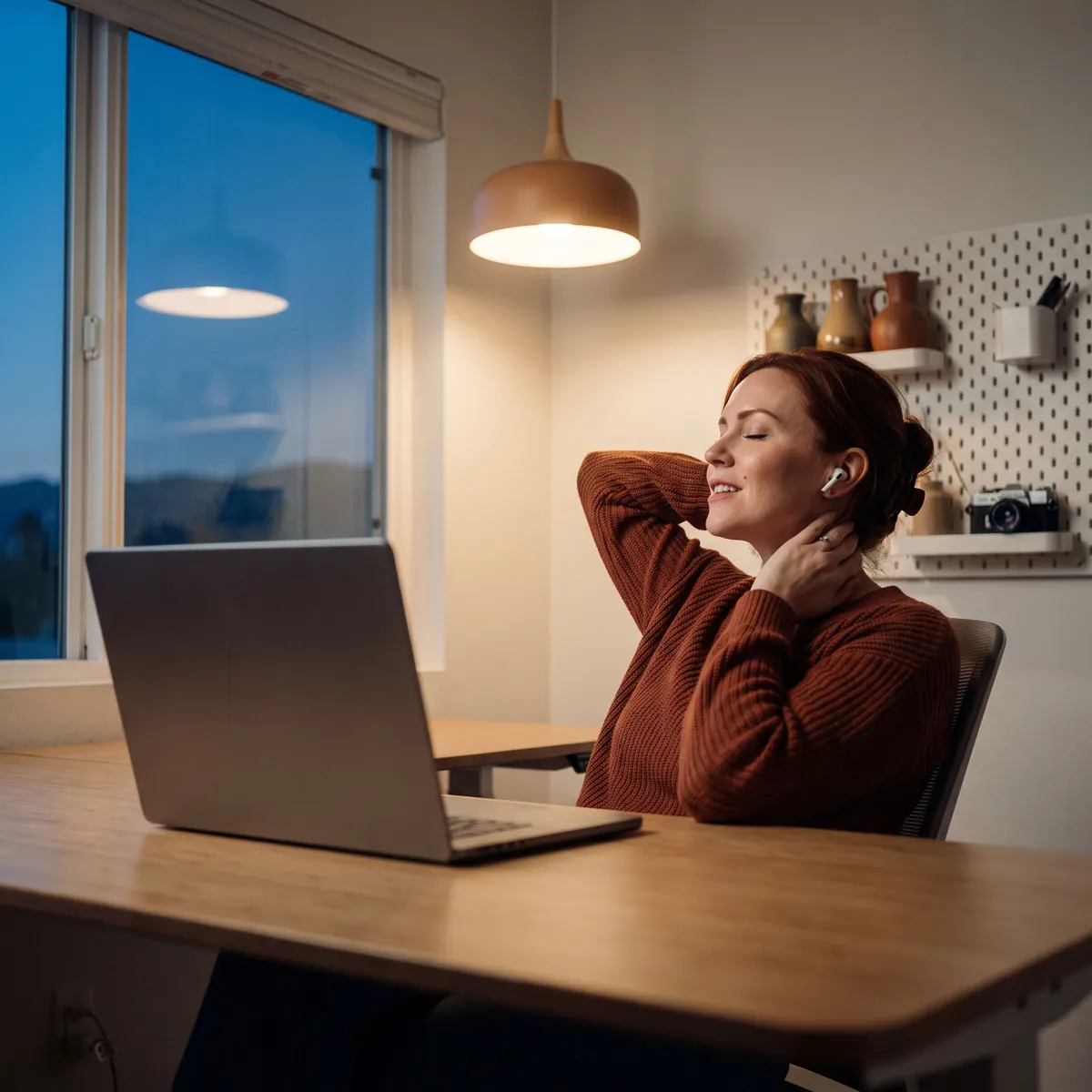 A designer leaning back at her desk in the evening, relieved, listening through earbuds